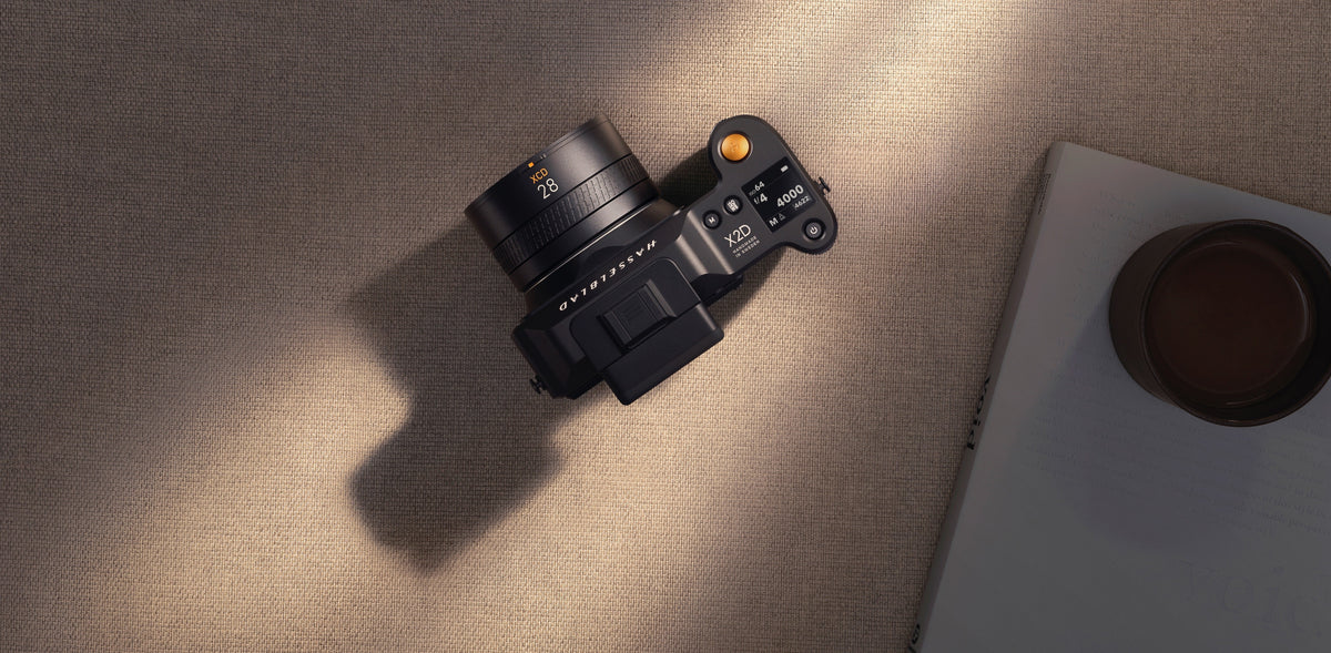 Hasselblad X2D 相機配 XCD 28 鏡頭，置於書本和杯子旁，在溫暖陽光下拍攝